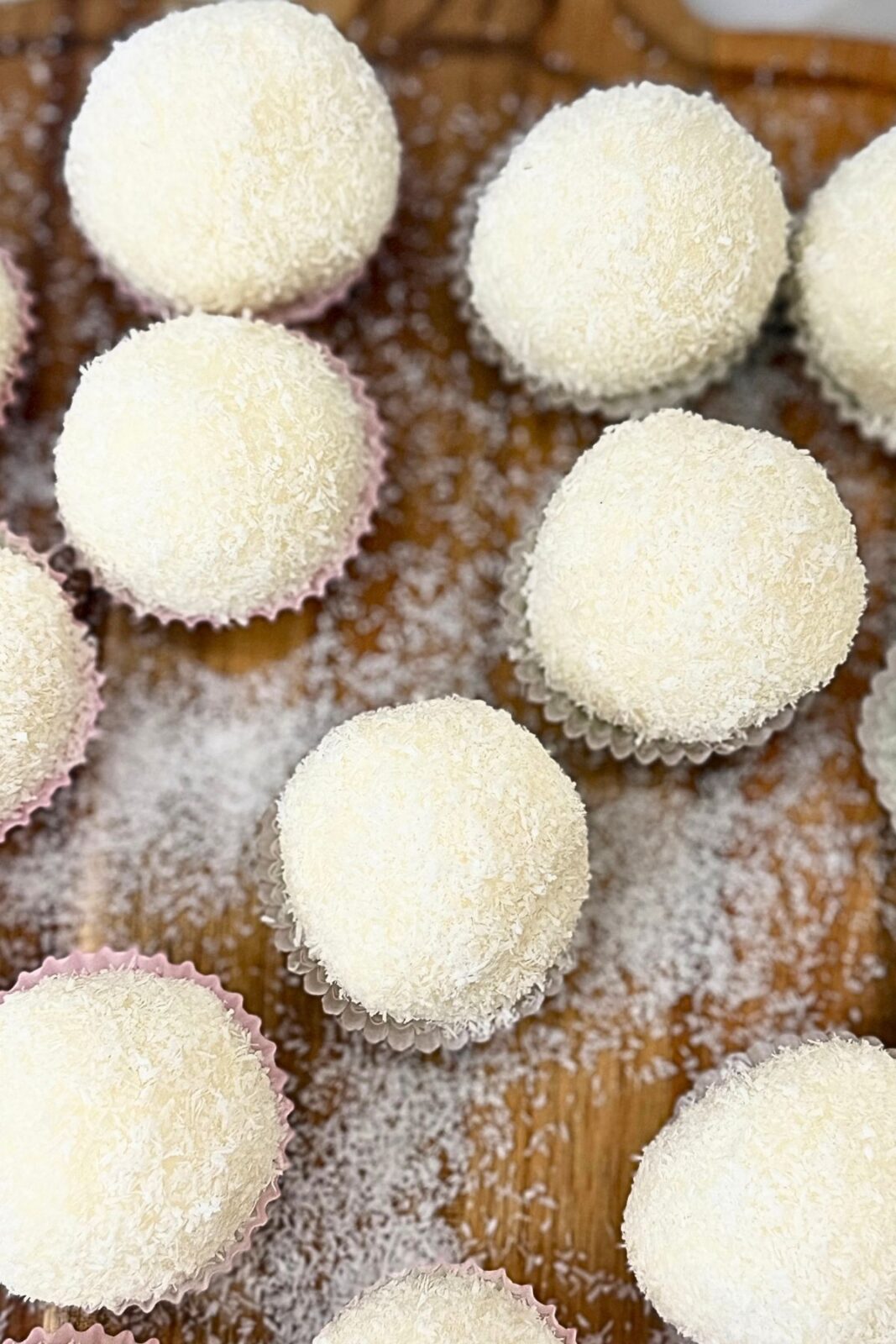 Finished Raffaello coconut balls coated in coconut and arranged in small paper cups on a wooden board.