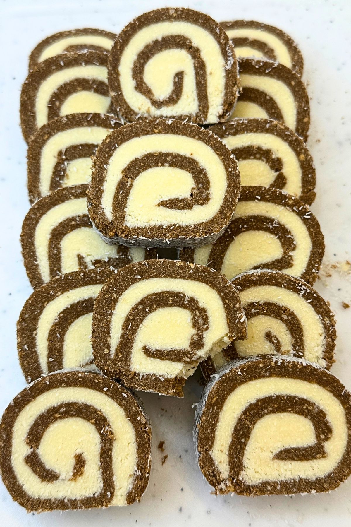 No-bake chocolate coconut roll slices stacked on a plate, showing cocoa cookie layers, white coconut filling, and a coconut coating.