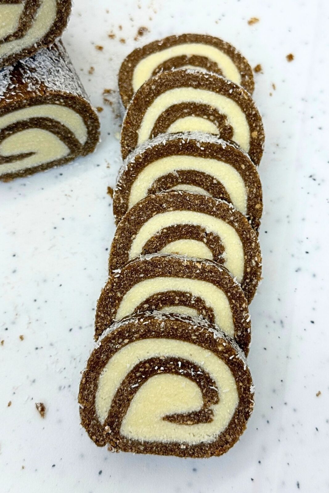 Chocolate coconut roll slices arranged in a row on a white platter, showing cocoa cookie spirals, coconut filling, and a coconut coating.