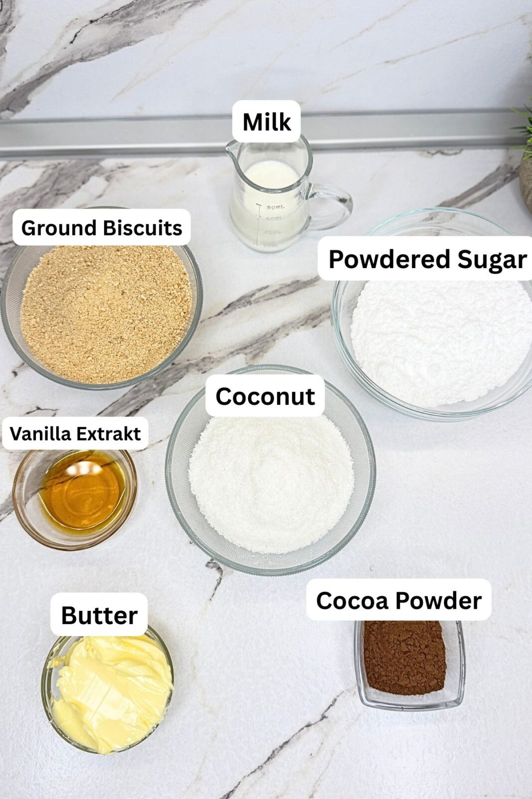 Ingredients for chocolate coconut roll, including cookie crumbs, powdered sugar, cocoa powder, milk, butter, and desiccated coconut on a marble countertop.