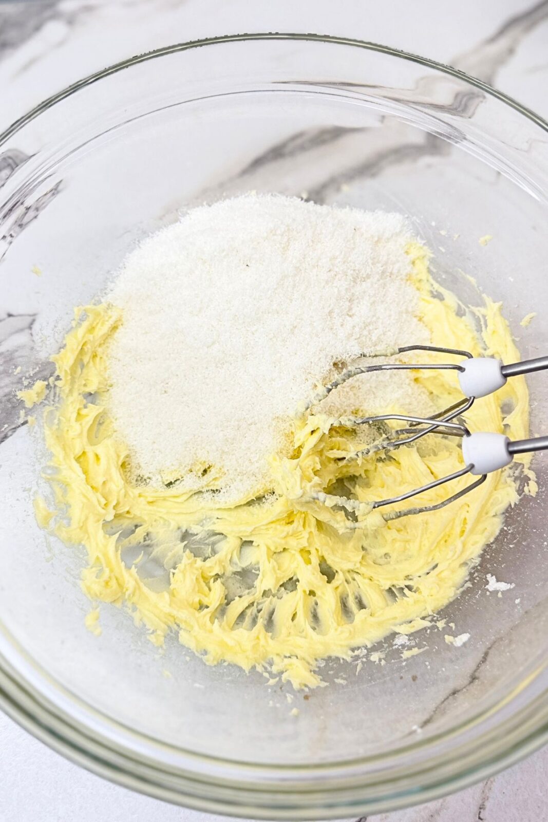 Desiccated coconut added to creamed butter and powdered sugar in a glass bowl with hand mixer beaters.