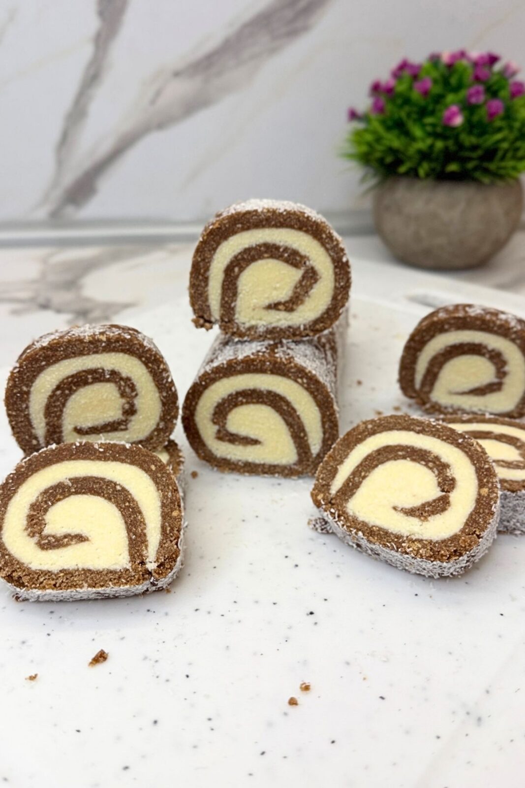 Chocolate coconut roll slices arranged on a white platter, with a slice in focus showing the cocoa cookie layer, coconut filling, and coconut-covered edge.