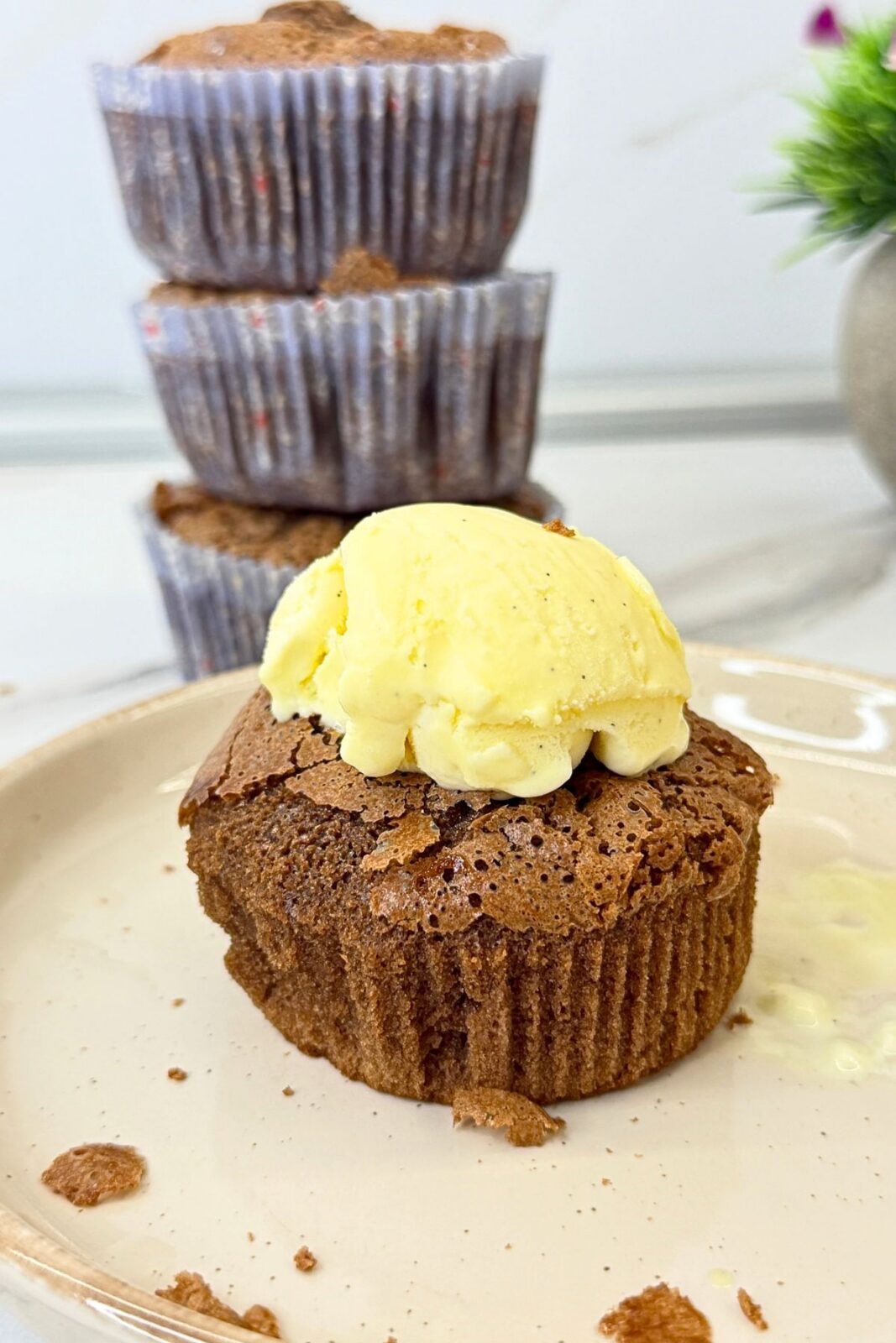 Chocolate lava cake topped with vanilla ice cream on a cream-colored plate, with three cakes in paper liners stacked in the background.