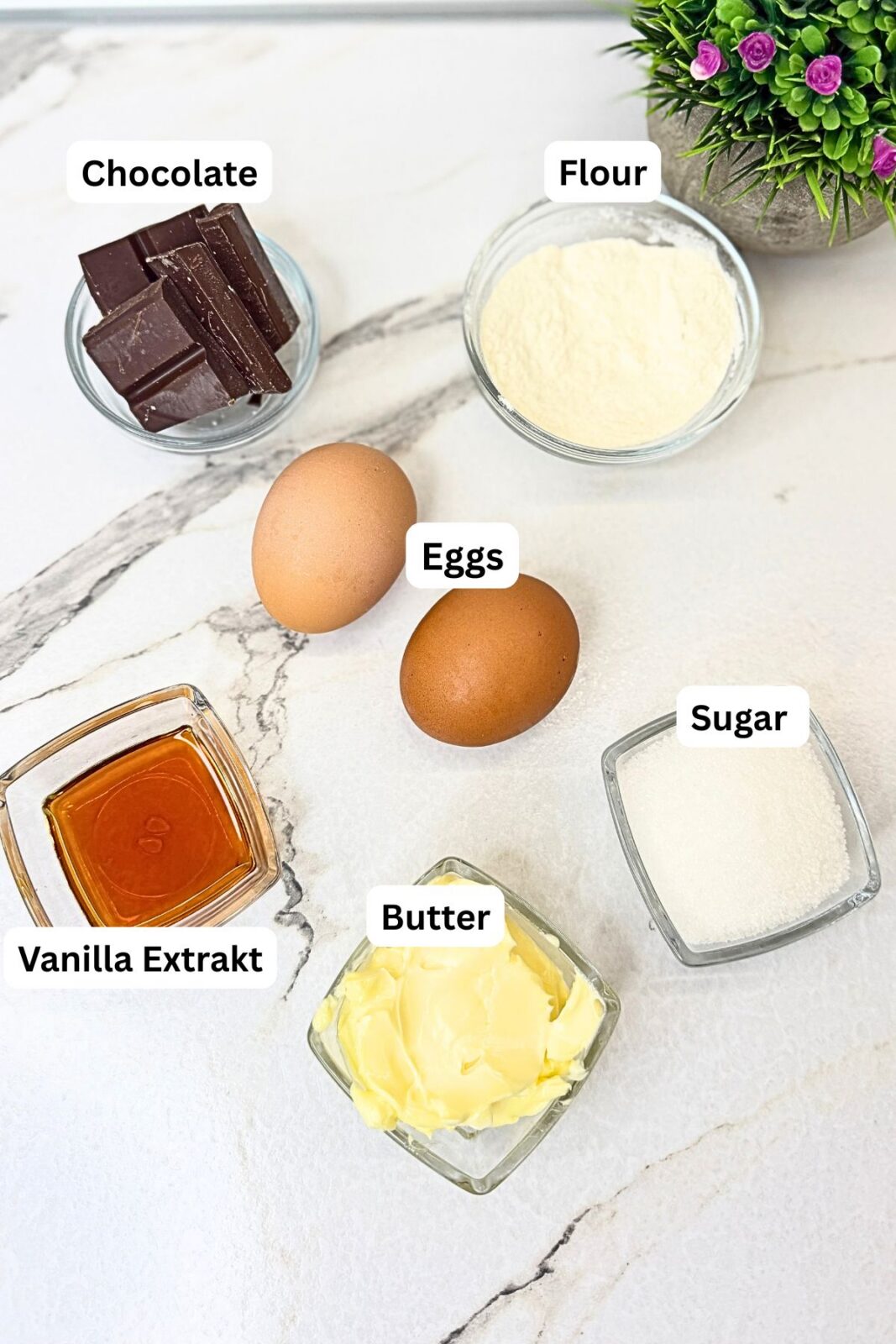 Ingredients for chocolate lava cakes: chocolate, flour, eggs, sugar, butter, and vanilla extract arranged on a marble countertop.