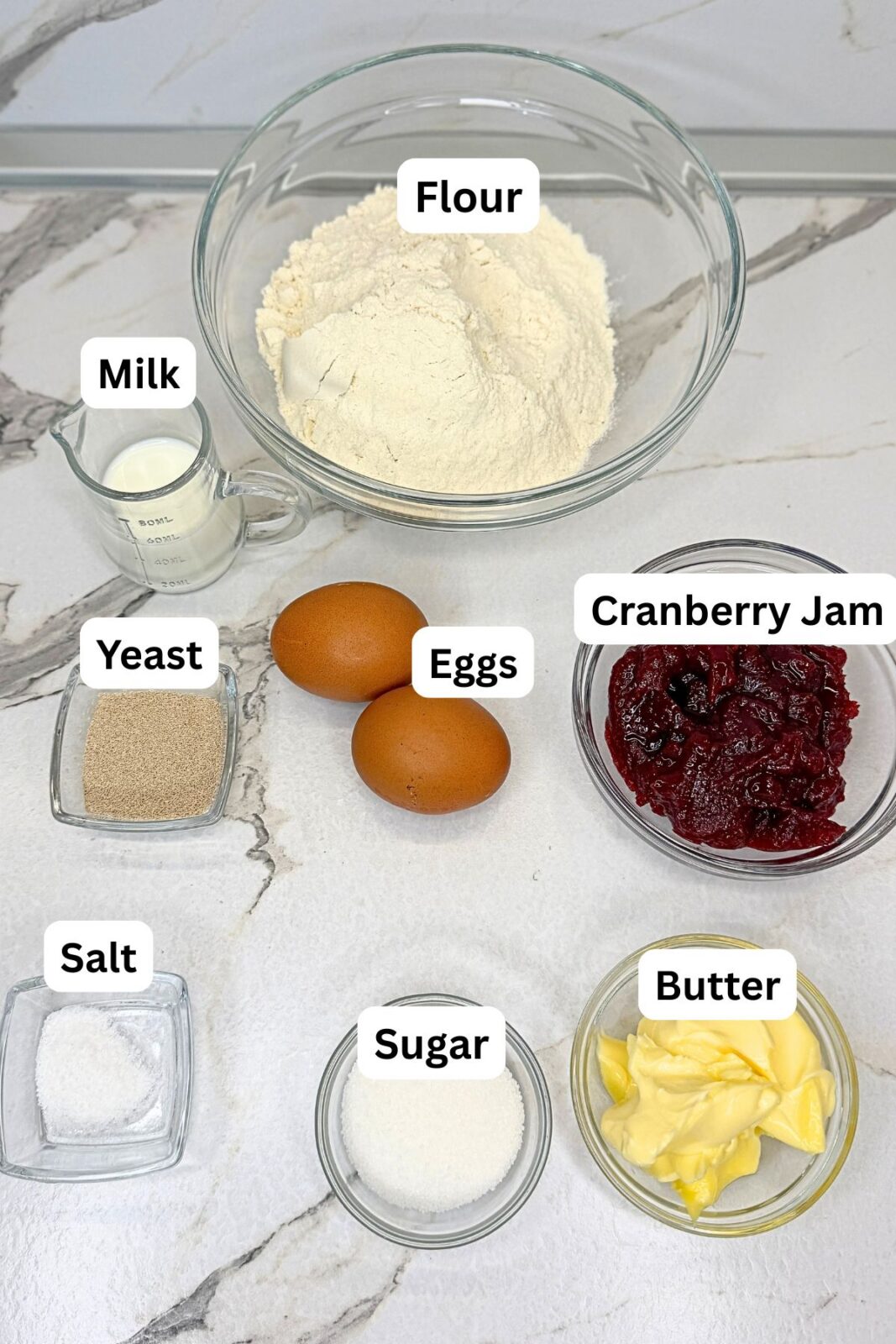 Ingredients for cranberry bread: flour, yeast, sugar, milk, butter, eggs, salt, and cranberry jam arranged on a marble countertop.