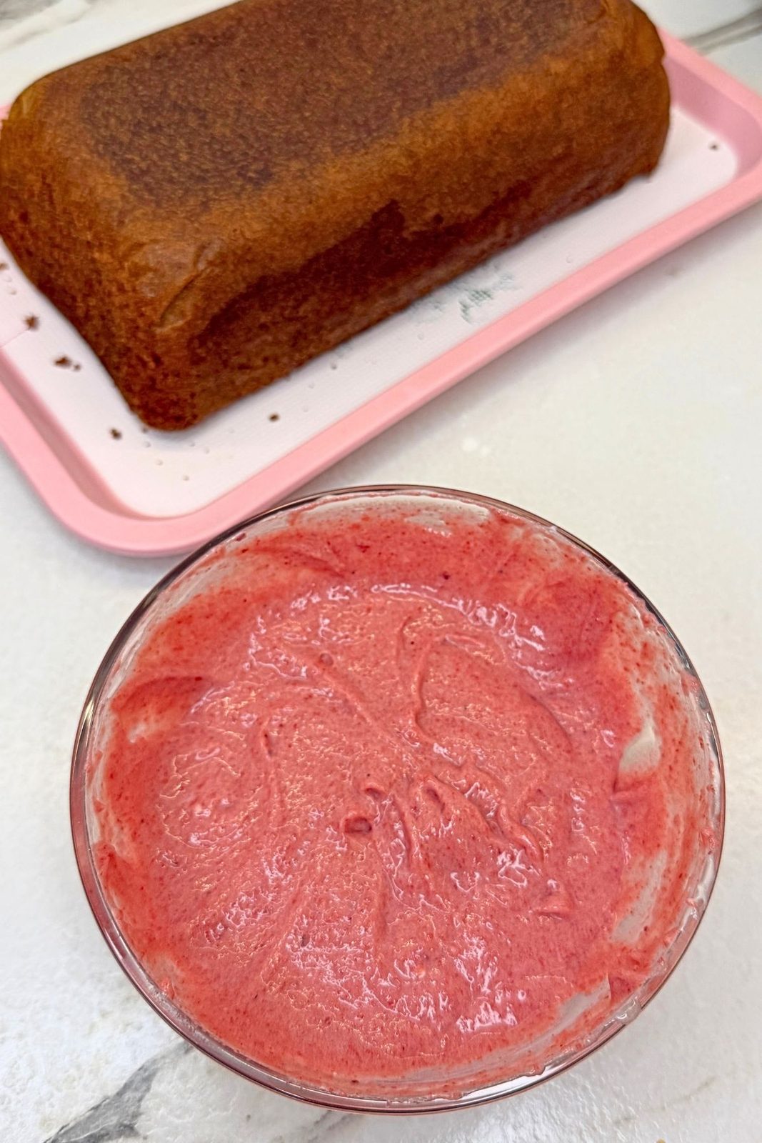 Baked cranberry loaf cake on a tray beside a glass bowl of pink cranberry cream cheese glaze.