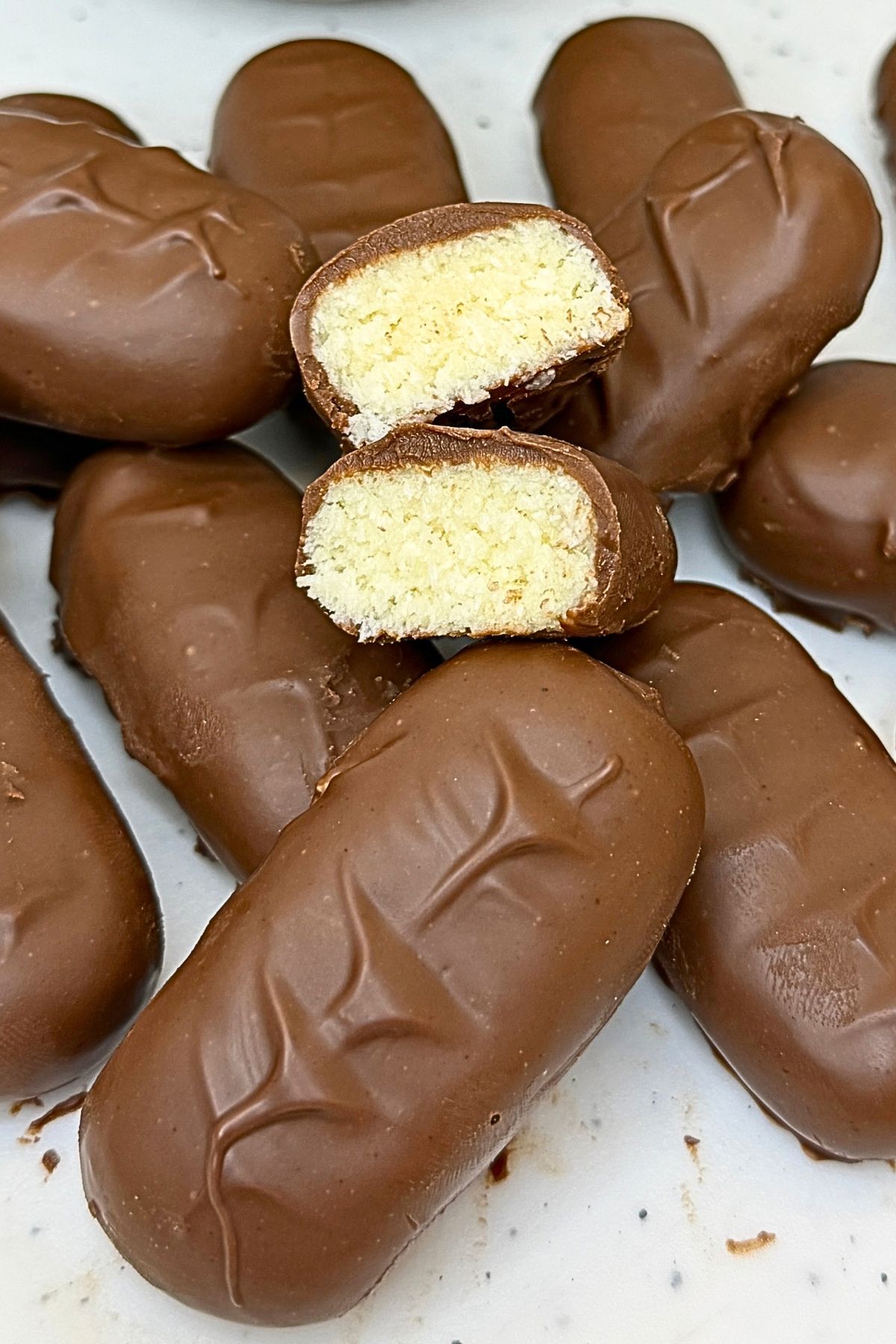 Homemade bounty bars coated in milk chocolate on a cream platter, with two cut halves showing the coconut filling.