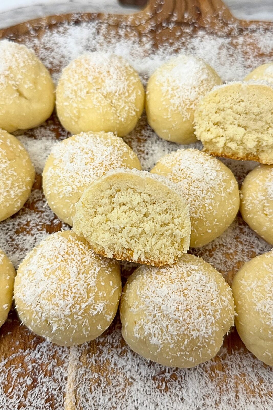 Lemon coconut shortbread cookies topped with lemon glaze and coconut, with a halved cookie showing the crumbly center on a wooden board.