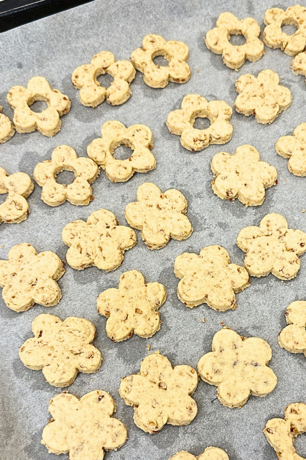 Unbaked flower-shaped Linzer cookie bases and cutout tops on a parchment-lined baking sheet.