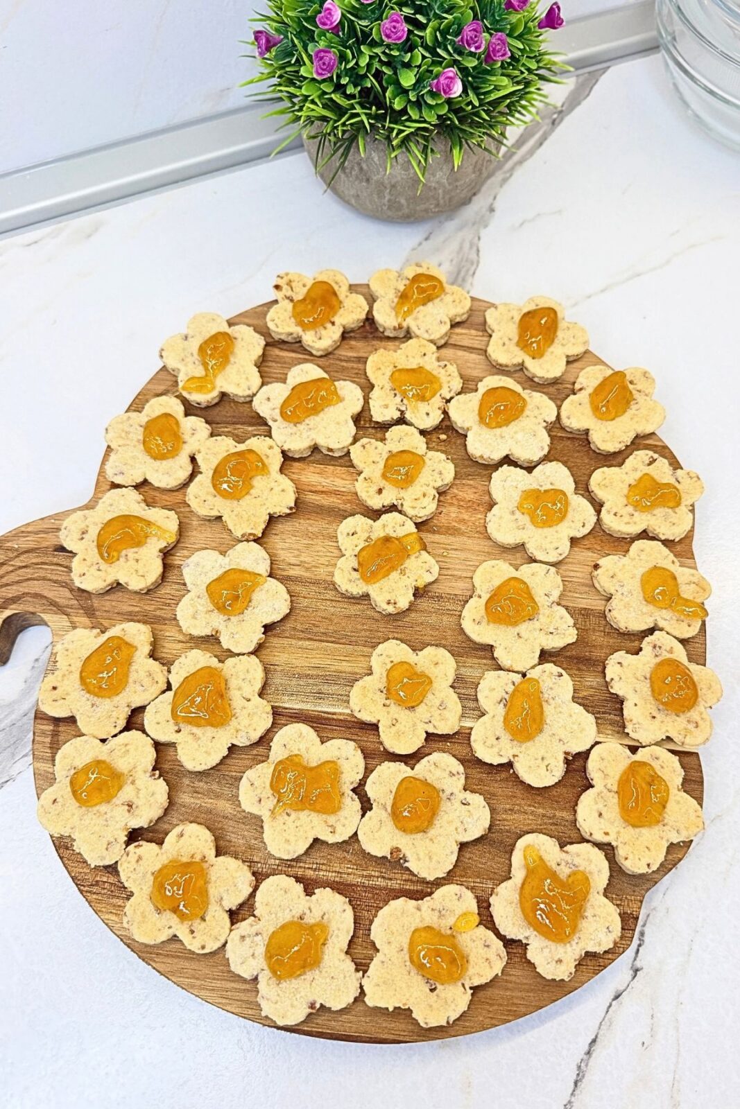 Flower-shaped almond cookie bases topped with apricot jam on a wooden board, ready for assembling Linzer cookies.