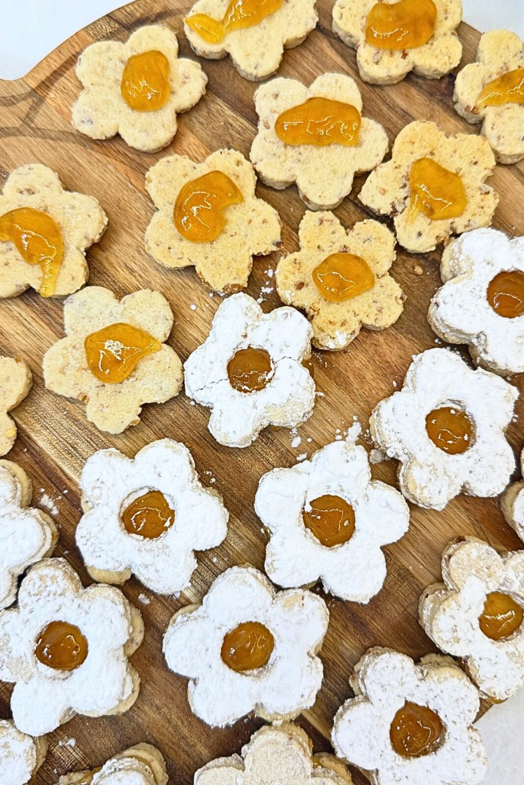 Flower-shaped Linzer cookies dusted with powdered sugar beside almond cookie bases topped with apricot jam on a wooden board.