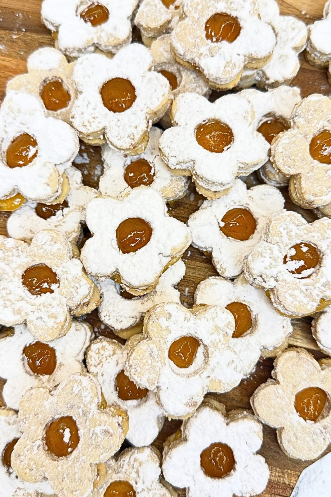 Close-up of flower-shaped Linzer cookies filled with apricot jam and dusted with powdered sugar, piled on a wooden board.