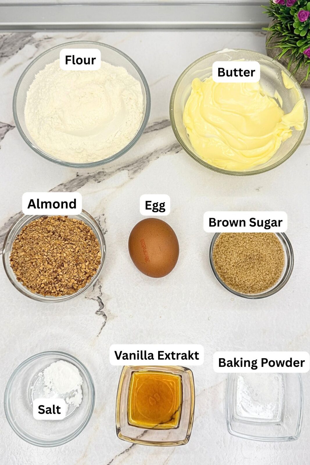 Ingredients for Linzer cookies arranged on a marble countertop: flour, butter, ground almonds, an egg, brown sugar, salt, vanilla extract, and baking powder.