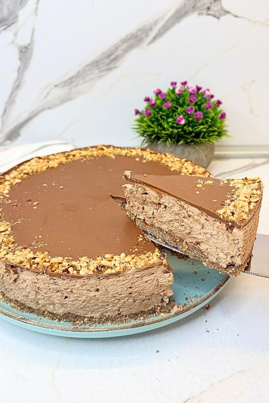 No-bake Ferrero Rocher cake with a slice lifted on a cake server, revealing the cocoa cookie crust, creamy Nutella filling, and chocolate topping edged with chopped hazelnuts.
