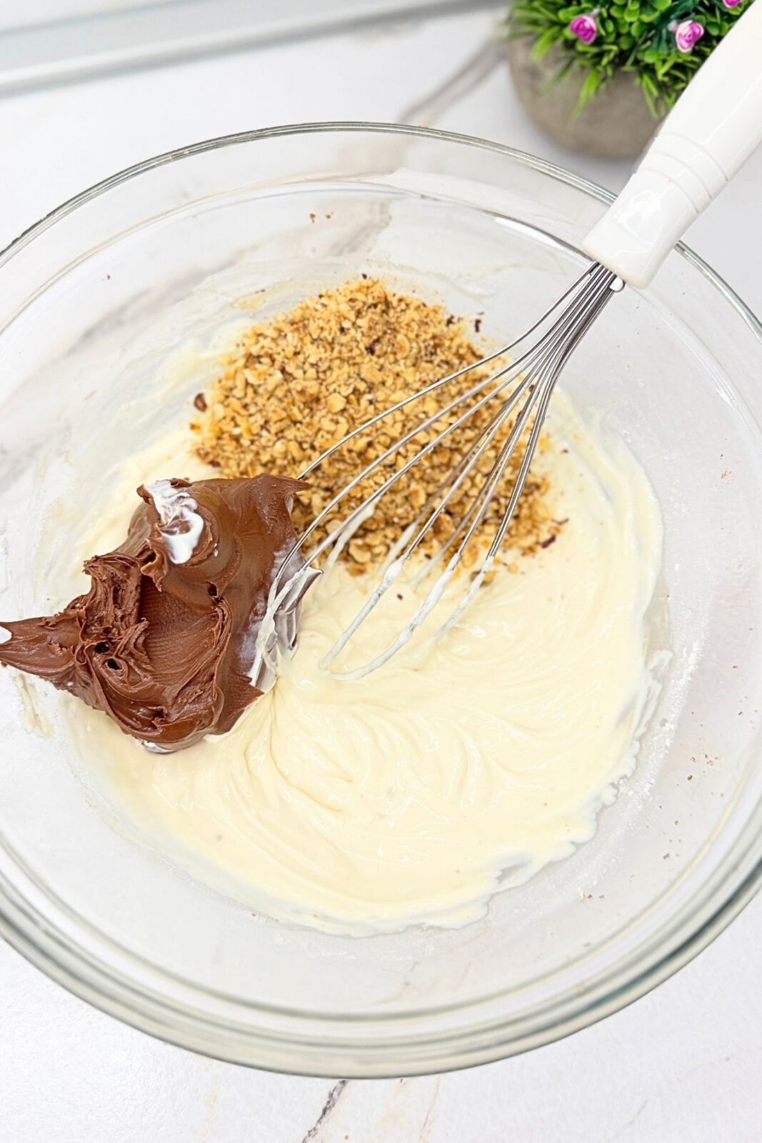 Nutella and ground hazelnuts added to a creamy mixture in a glass bowl for no-bake Ferrero Rocher cake.