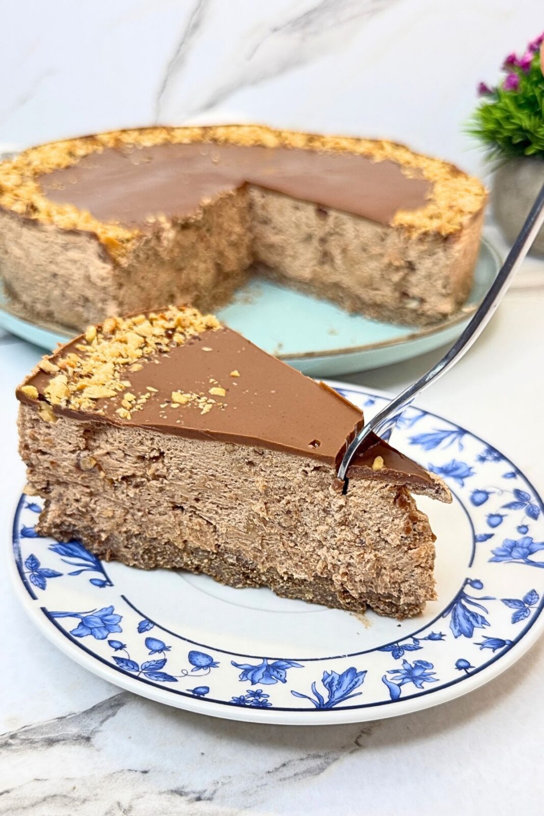 Slice of no-bake Ferrero Rocher cake on a blue-and-white plate, showing the cocoa cookie crust, creamy Nutella filling, chocolate topping, and chopped hazelnuts.