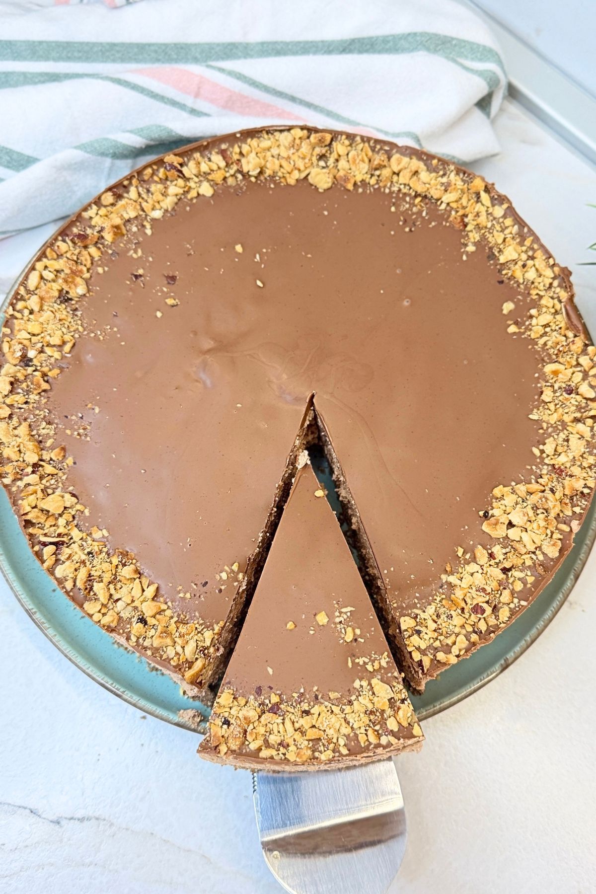 No-bake Ferrero Rocher cake with a slice lifted on a cake server, showing the cookie crust, creamy Nutella filling, chocolate topping, and chopped hazelnuts.