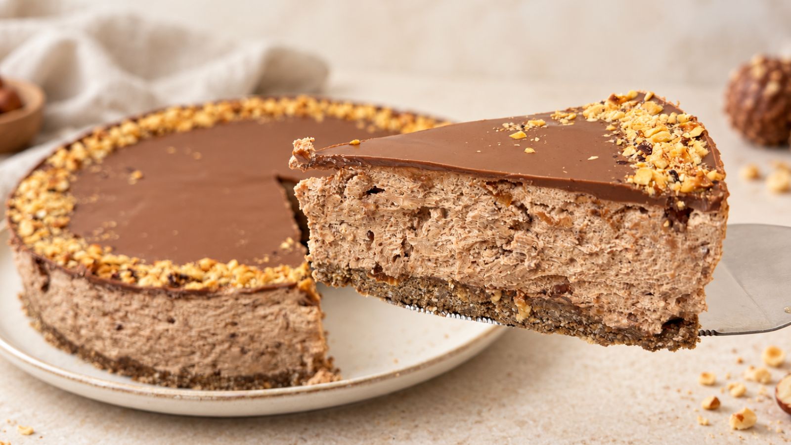 No-bake Ferrero Rocher cake with a slice lifted on a cake server, showing the cookie crust, creamy Nutella filling, chocolate topping, and chopped hazelnuts.