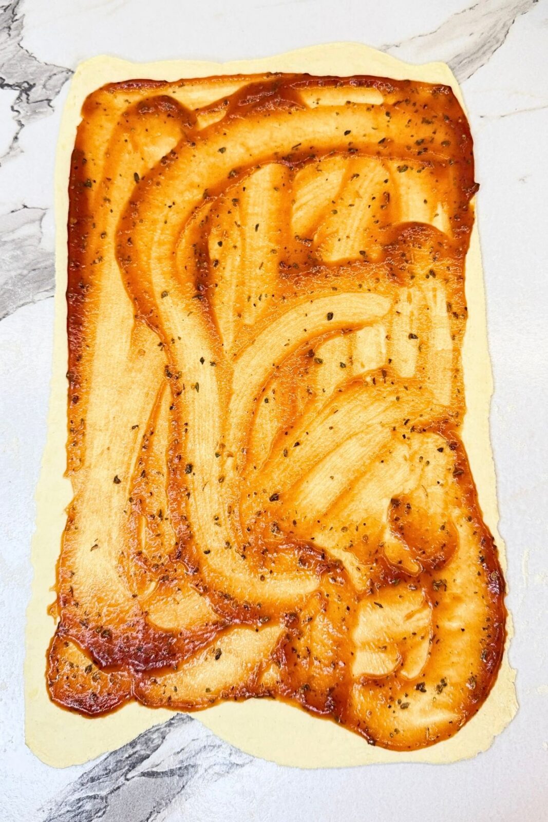 Puff pastry spread with pizza sauce on a lightly floured marble countertop.