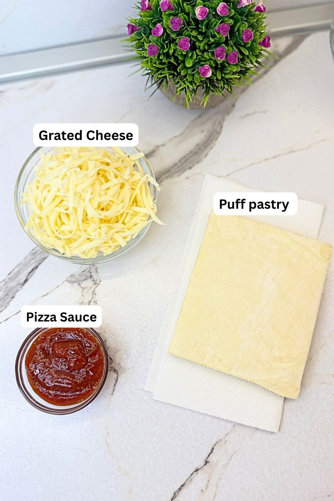 Ingredients for pizza rolls: puff pastry, shredded cheese, and pizza sauce arranged on a marble countertop.