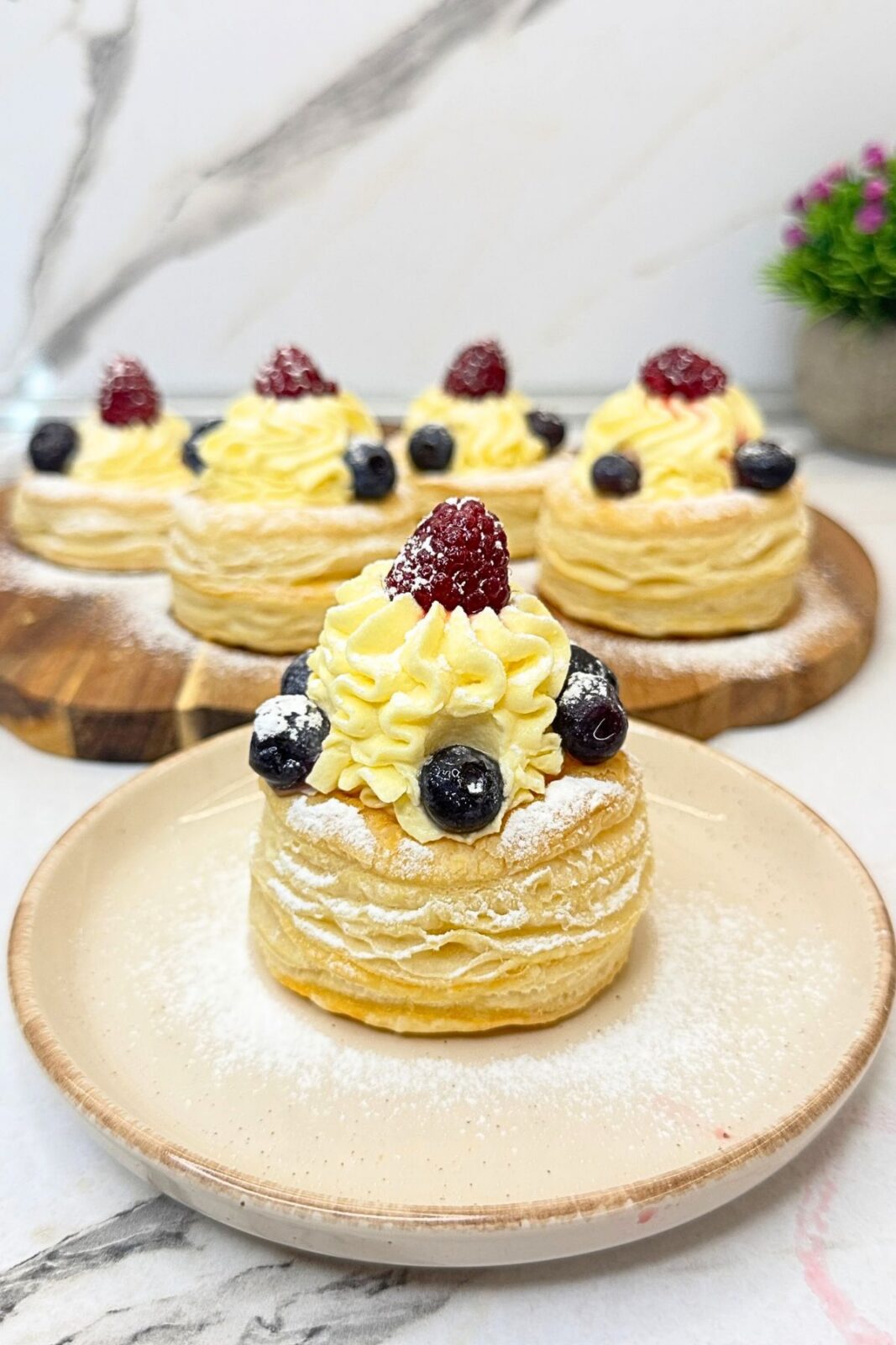 Puff pastry cups with piped vanilla cream, blueberries, raspberries, and powdered sugar, with one served on a small plate.