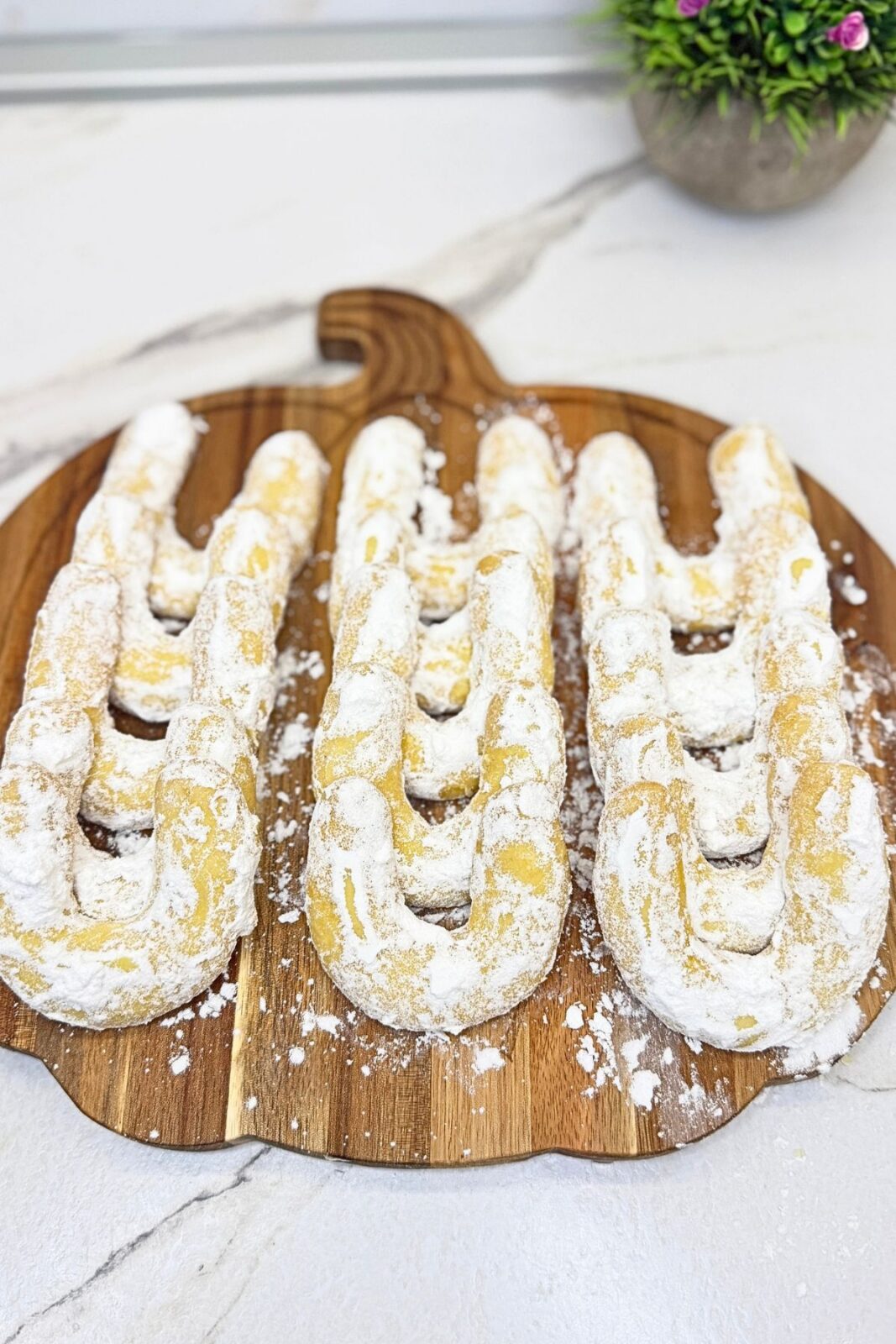 Vanilla crescent cookies coated in powdered sugar and arranged in rows on a wooden serving board, with flowers in the background.