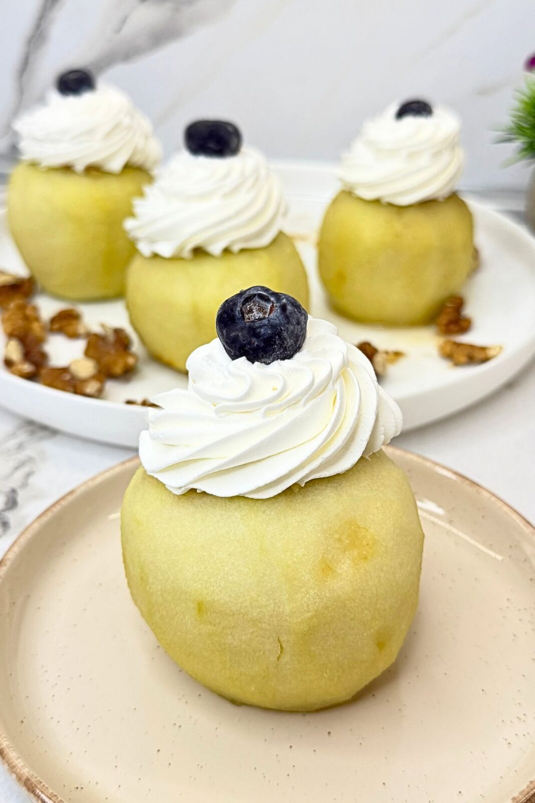 Walnut stuffed apple topped with whipped cream and a blueberry on a beige plate, with more stuffed apples in the background.