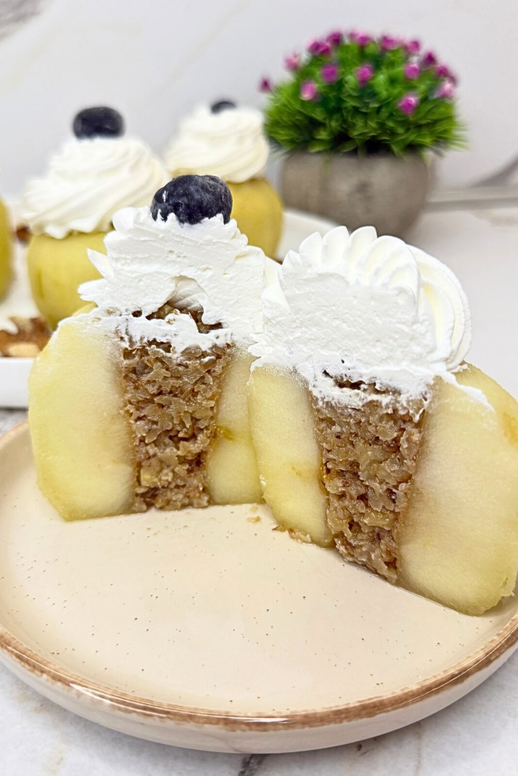 Walnut stuffed apple cut in half to show the walnut filling, topped with whipped cream and a blueberry on a beige plate.
