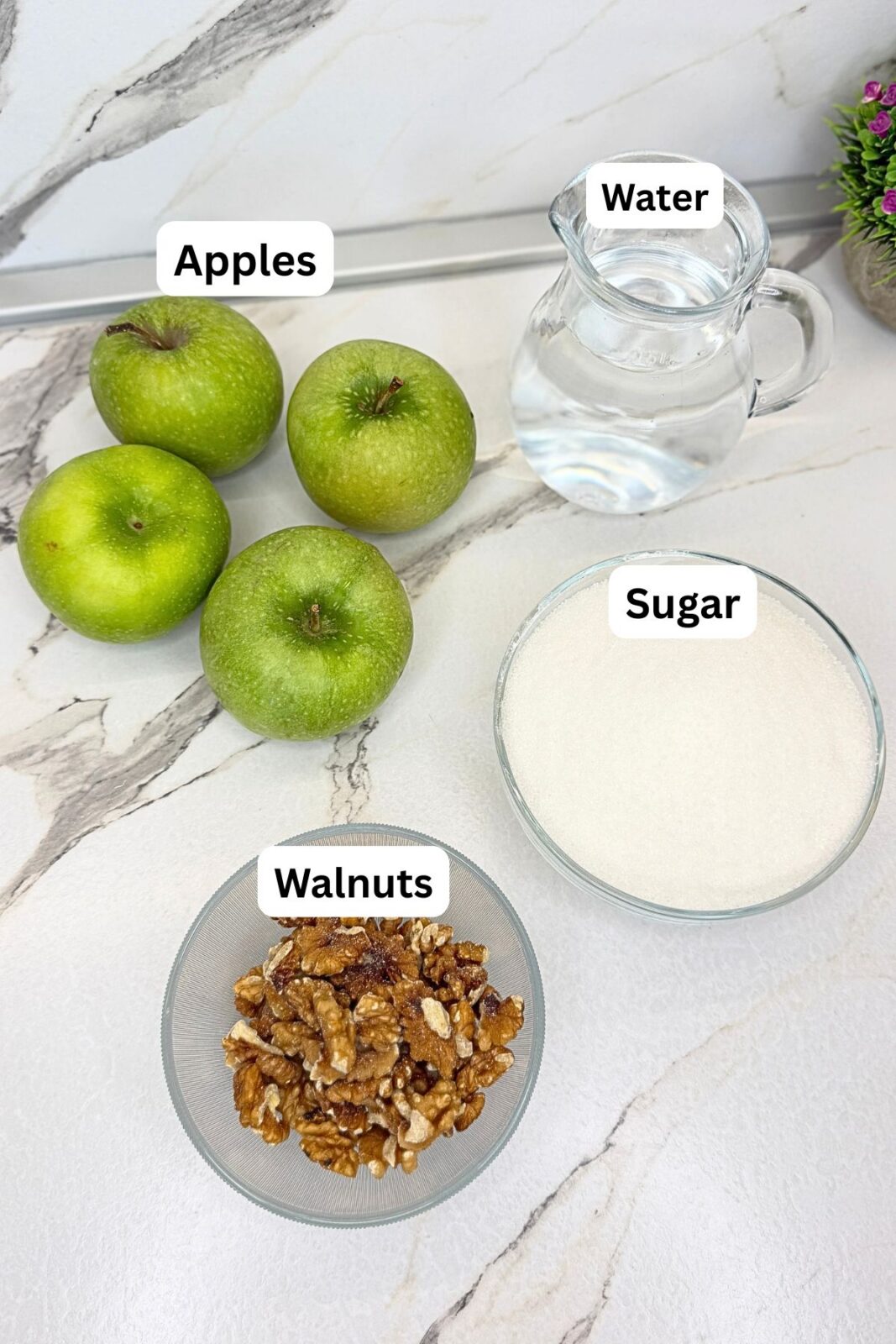 Ingredients for walnut stuffed apples: Granny Smith apples, water, granulated sugar, and walnuts arranged on a marble countertop.